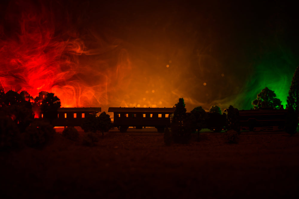 A dark passenger train at night, windows glowing orange, surrounded by trees and thick red and green smoke.