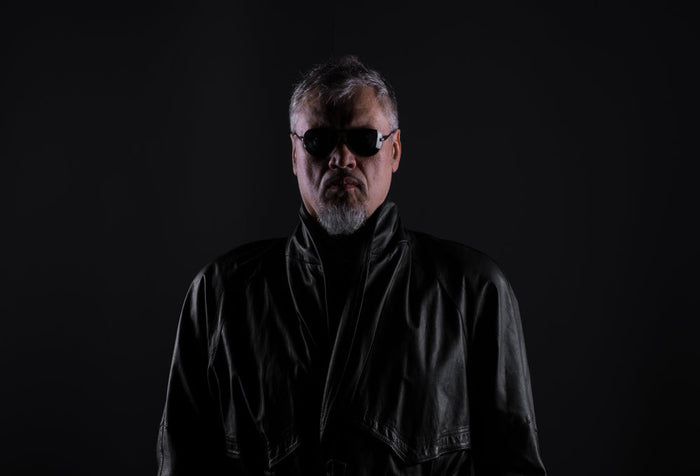 A man with gray hair and a goatee stands against a black background, wearing dark sunglasses and a black leather coat with the collar turned up, face lit dimly.