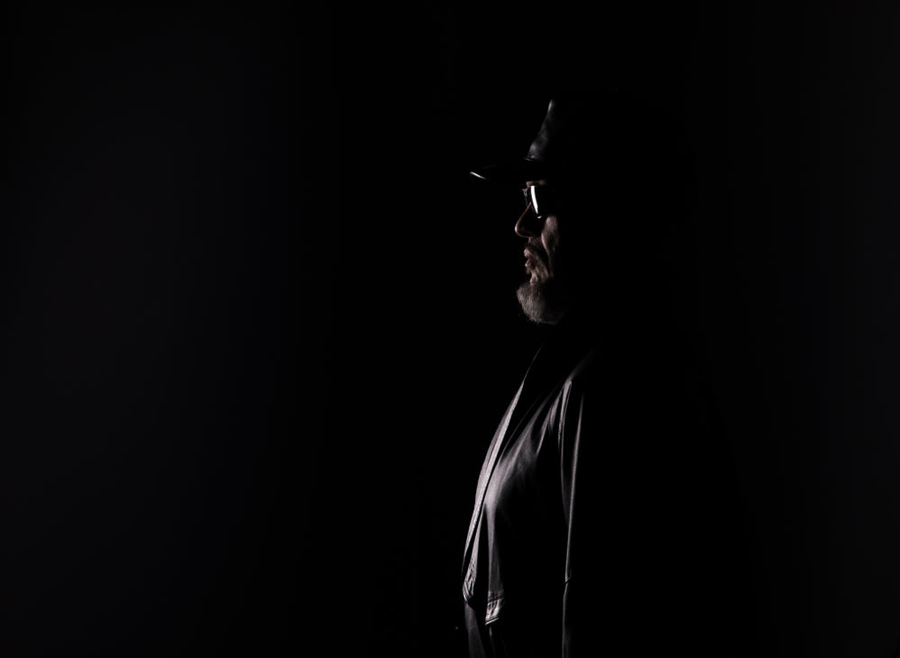 An elderly man in profile, lit from the front against a black background, wearing a cap, glasses, and a jacket, his face mostly in shadow.