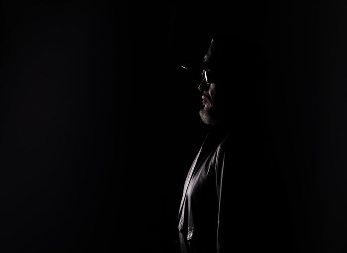 An elderly man in profile, lit from the front against a black background, wearing a cap, glasses, and a jacket, his face mostly in shadow.