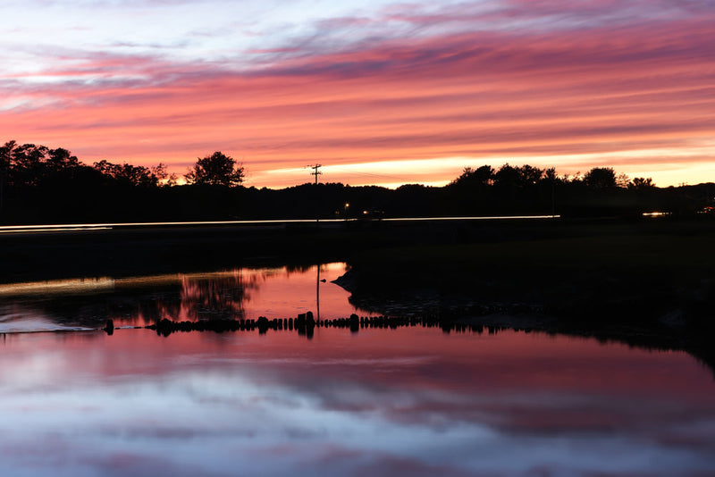 Still water at dusk reflecting a pink and purple sky, with a dark treeline and old wooden pilings silhouetted across the far bank.