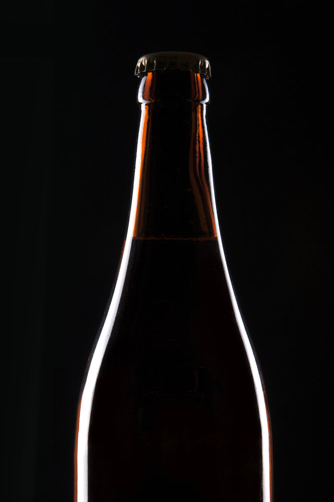 The neck and shoulder of a capped brown glass beer bottle, rim-lit against a black background.