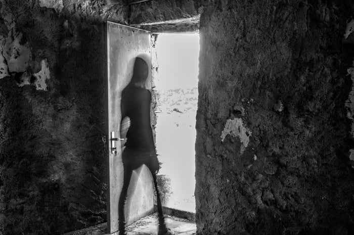 A shadowy figure slips through an open door into harsh daylight, leaving a dark, decaying stone room in an eerie black-and-white scene.