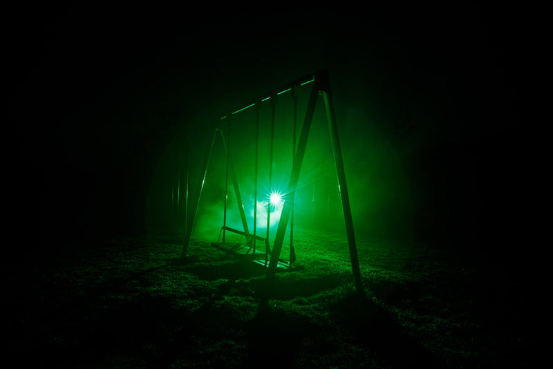 An empty porch-style swing on a wooden frame at night, backlit by a bright green glow and low fog on the grass.