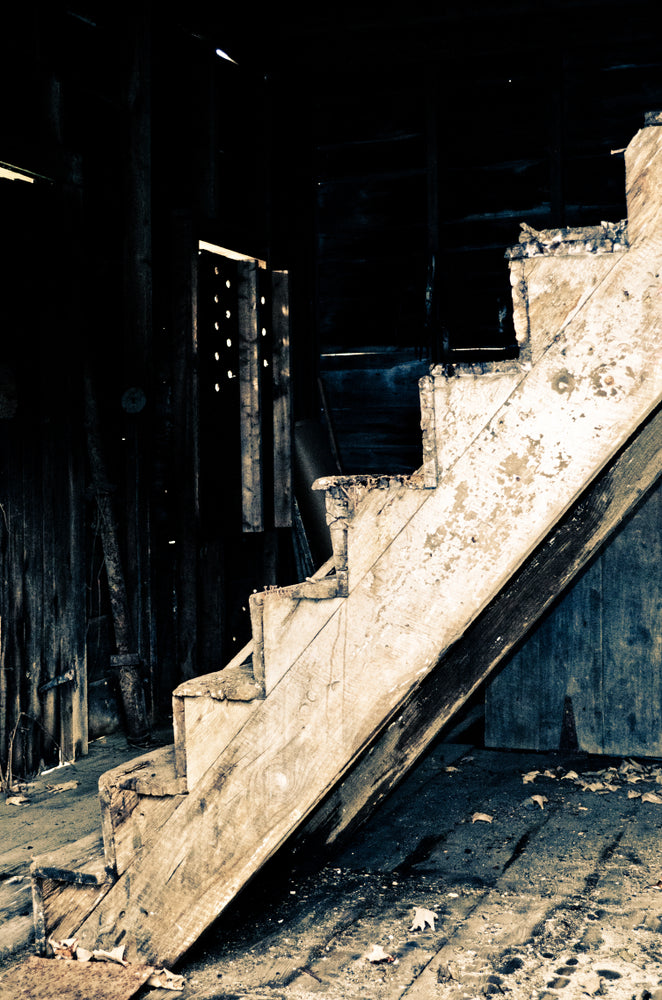 A worn wooden staircase inside a dark, derelict building, its steps caked in dust and grime, with debris scattered across the floor below.