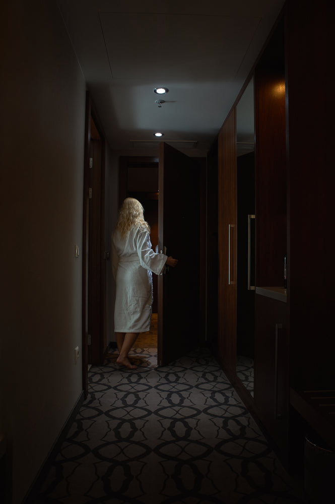 A woman in a white robe stepping through a doorway into a dark room at the end of a dim hallway, seen from behind.