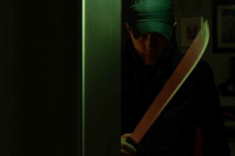 A man in a dark cap and jacket grips a machete while lurking behind a doorway in a dim, green-lit room.