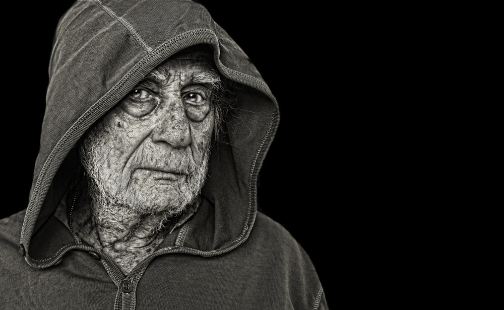 An elderly homeless man in a worn hooded jacket, his deeply lined face turned toward the light against a black background.
