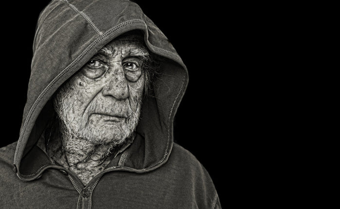 An elderly homeless man in a worn hooded jacket, his deeply lined face turned toward the light against a black background.