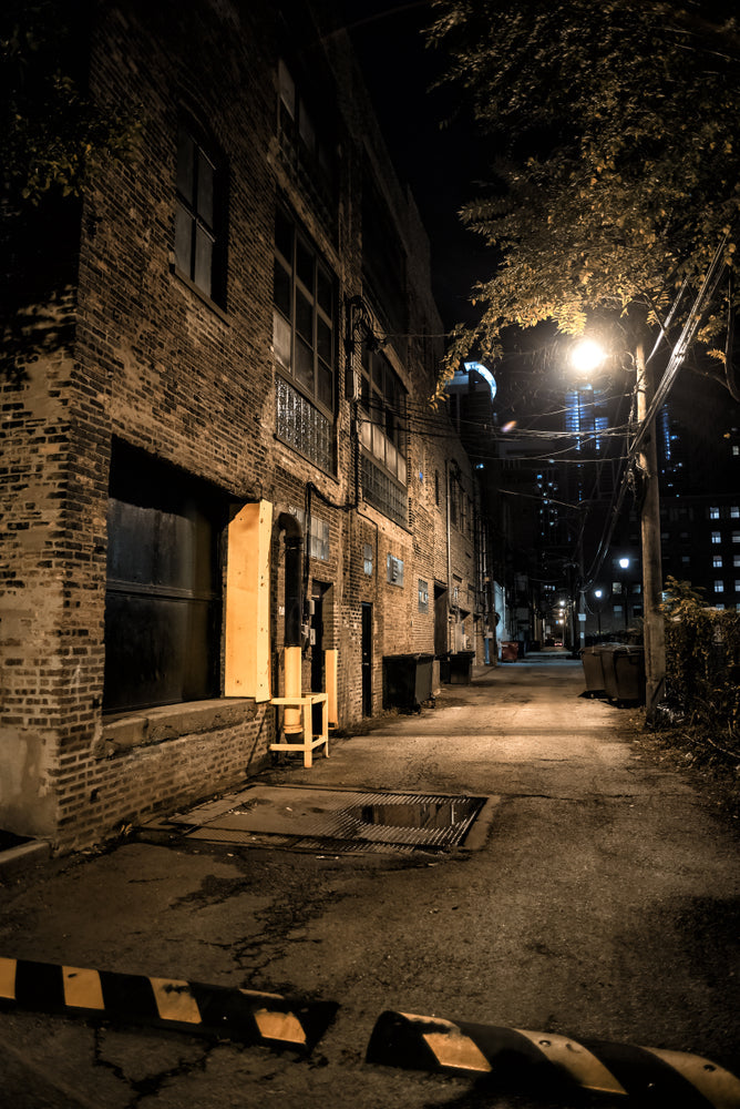 A dark, empty downtown alley at night, lit by a single street lamp, lined with old brick buildings, dumpsters, and a distant lit high-rise.