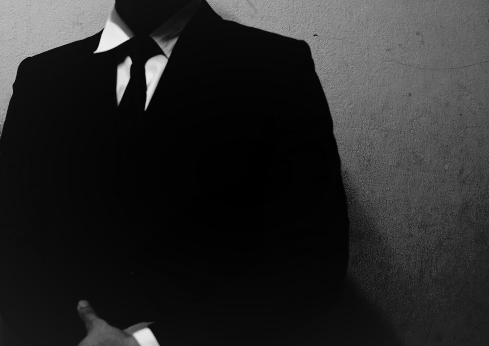 Black-and-white image of a faceless man in a dark suit and tie standing against a bare wall, his head cropped from the frame.