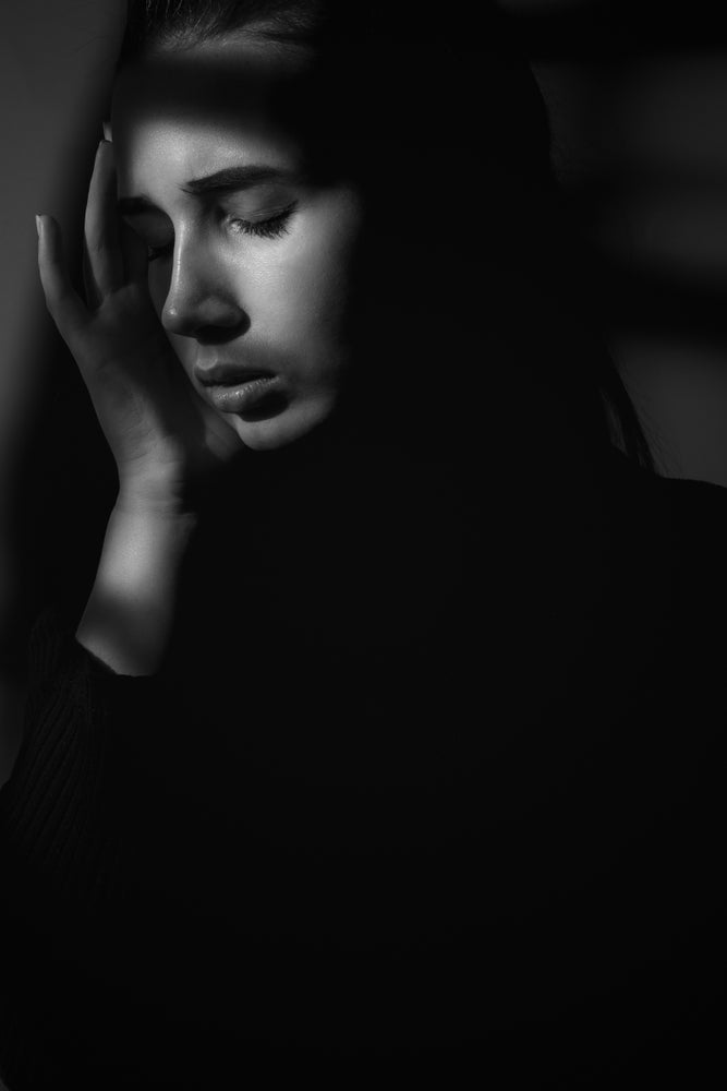 Black-and-white portrait of a woman with closed eyes resting her face in one hand, partially obscured by deep shadows across her face and body.