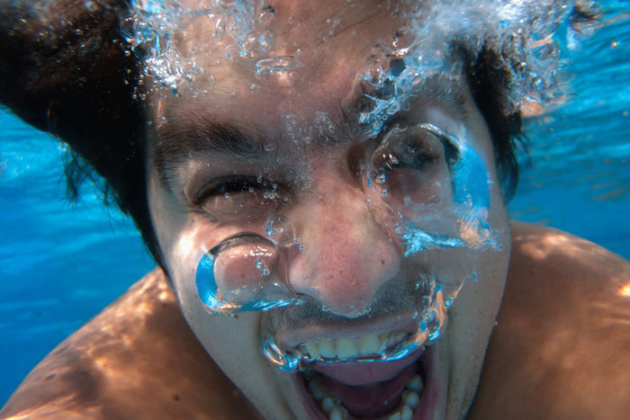 A man's face underwater in bright blue water, mouth open with air bubbles streaming past his face.