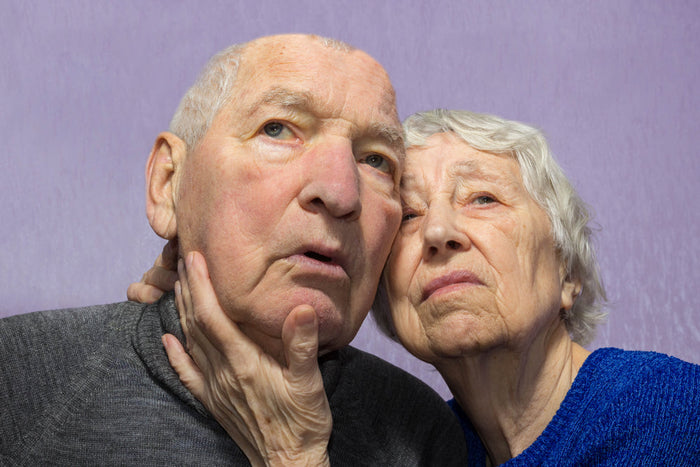 Elderly couple posed closely together, the woman gently holding the man's face as both look upward against a plain lavender background.