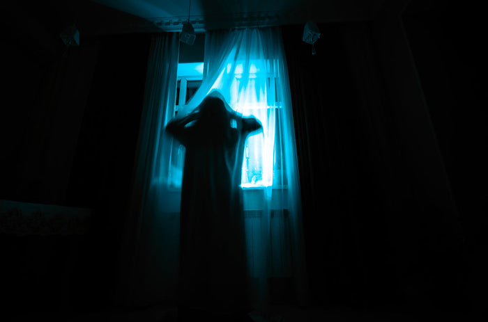 A shadowy figure in a pale gown standing at a curtained window at night, backlit by glowing blue light, hands raised to its head, face obscured.