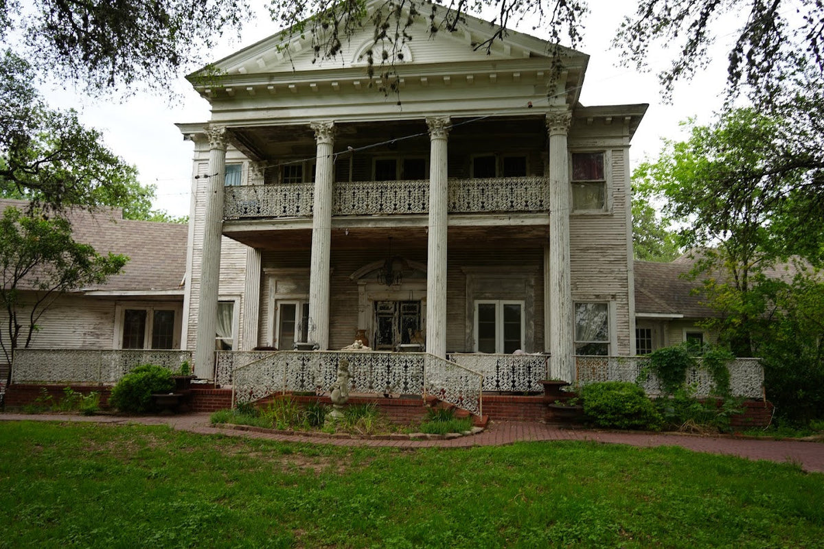 Weathered two-story Southern mansion with tall white columns, ornate iron railings, broad porches, and overgrown greenery beneath oak trees.