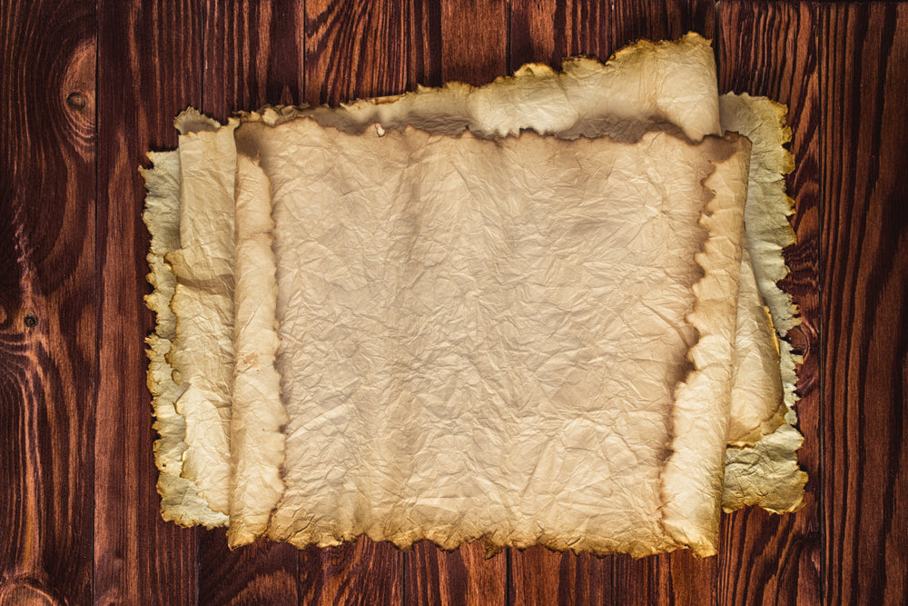 Stack of aged, wrinkled parchment sheets with torn, darkened edges resting on a richly grained wooden tabletop.
