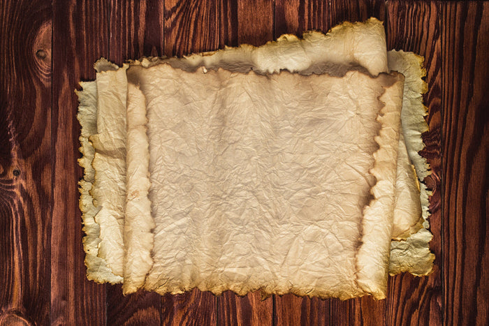 Stack of aged, wrinkled parchment sheets with torn, darkened edges resting on a richly grained wooden tabletop.