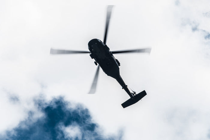 Helicopter viewed from below in dark silhouette, its rotor blades blurred in motion against a bright, cloudy blue-and-white sky.