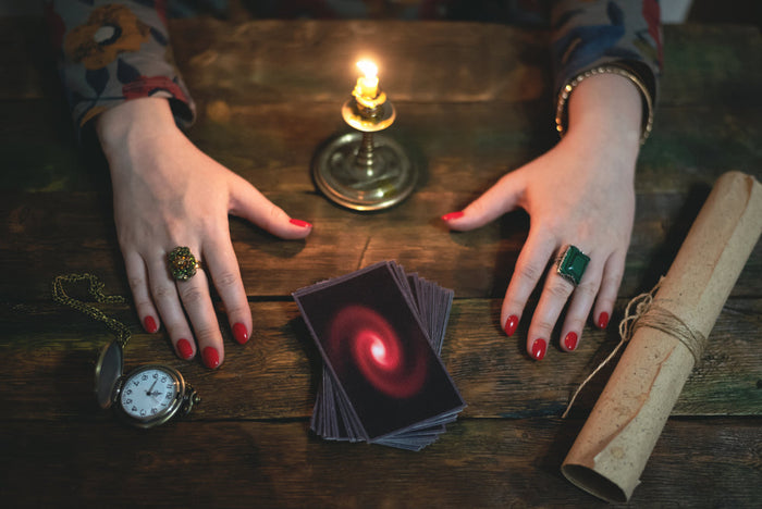 A fortune-teller's hands resting on a rustic wooden table lit by a single candle, with a deck of face-down cards, a pocket watch, and a rolled scroll.