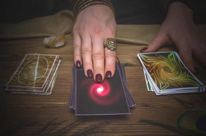 A fortune teller’s hands hover over three decks of cards on a wooden table, with a glowing red symbol illuminating the center deck.