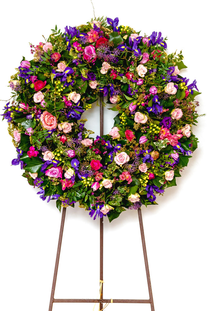Large funeral wreath overflowing with pink roses, purple flowers, greenery, and berries, displayed on a simple metal stand.