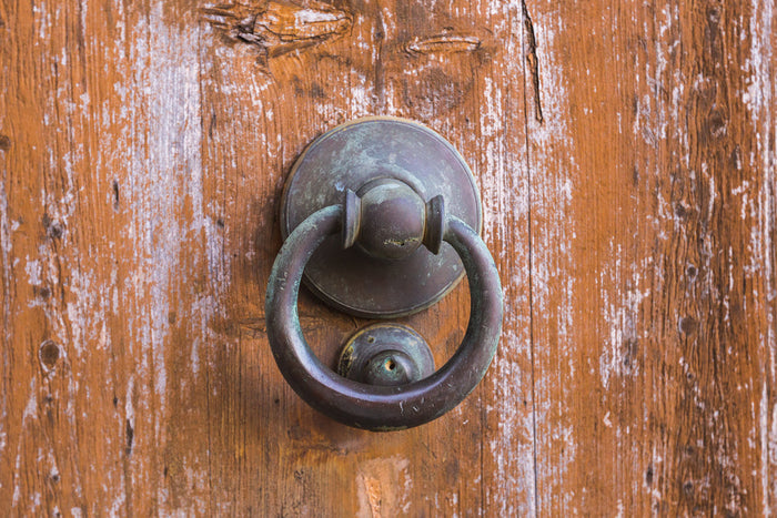 A weathered bronze ring door knocker mounted on an aged wooden door with peeling white paint, suggesting an old, seldom-used entrance.