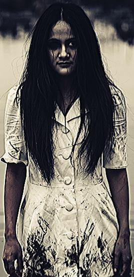 A pale, ghostly woman with long black hair and a stained white dress stands rigidly against a dark, blurred background.