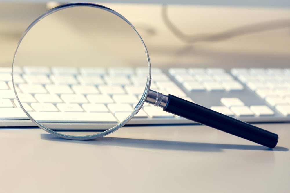 A magnifying glass rests in front of a computer keyboard, symbolizing investigation, research, or a search for hidden information.