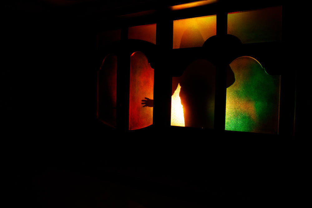 A shadowy figure reaches from behind stained glass panels glowing red, orange, and green, creating an ominous scene against deep darkness.