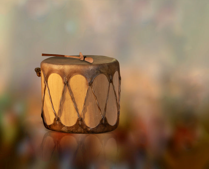 A traditional hand drum with a hide-covered top and wooden beater rests against a softly blurred background in warm, earthy tones.