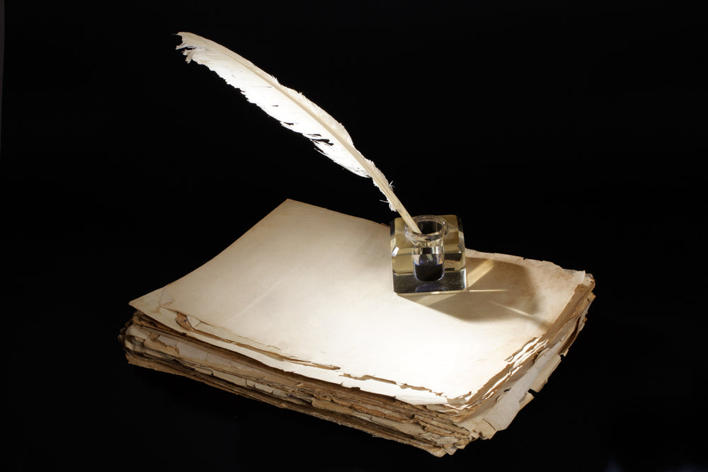 White feather quill resting in a glass inkwell atop a stack of aged, worn parchment pages against a dramatic black background.