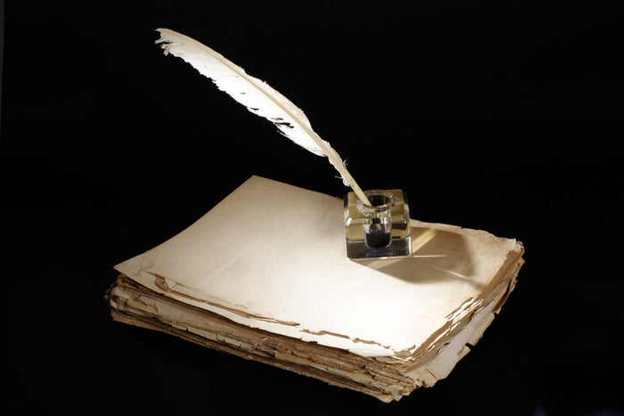 White feather quill resting in a glass inkwell atop a stack of aged, worn parchment pages against a dramatic black background.